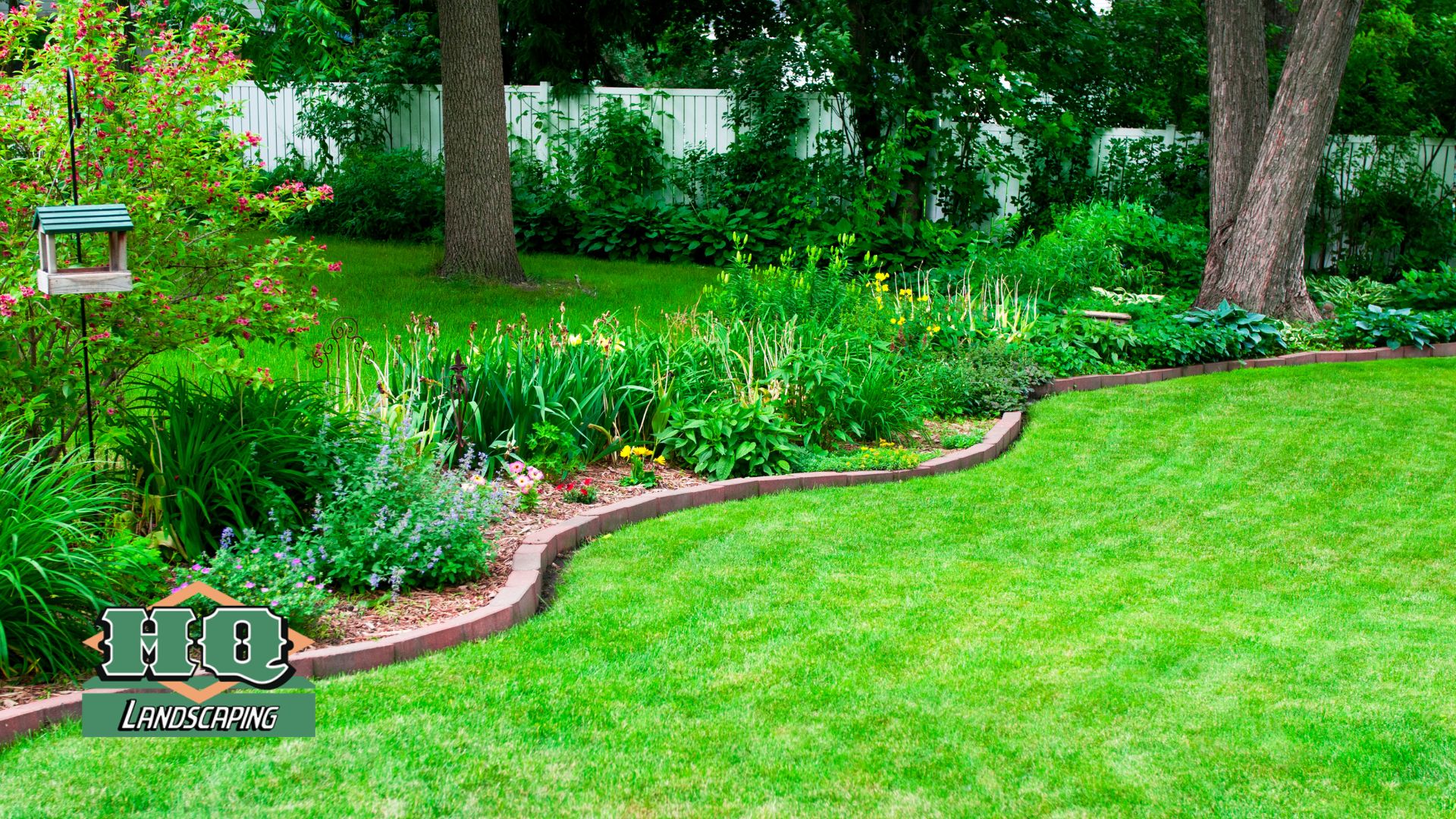 A beautifully maintained green lawn with a curved flower bed, colorful plants, and a wooden bird feeder, showcasing HQ Landscaping's professional lawn installation and landscaping services.